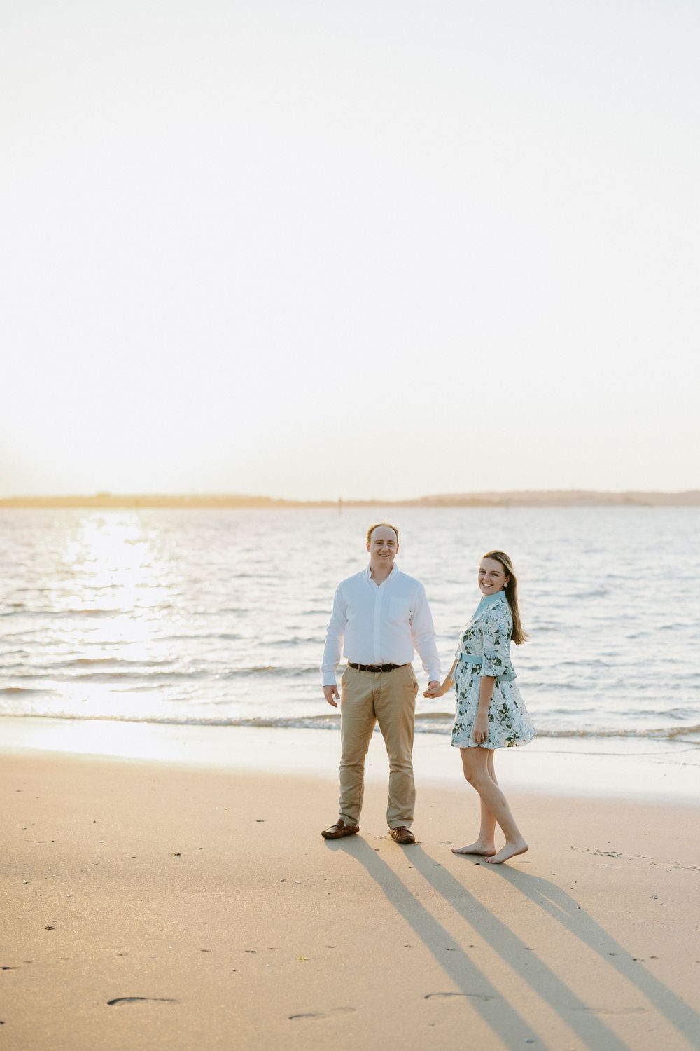Couple holding hands on sandy beach at golden hour, man in white shirt and woman in floral dress smiling at camera.