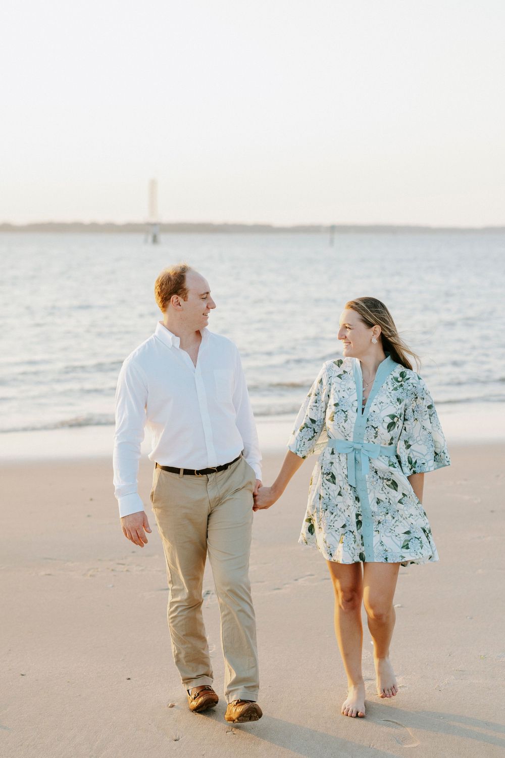 Couple holding hands and walking on a sandy beach, man in white shirt and woman in floral dress.