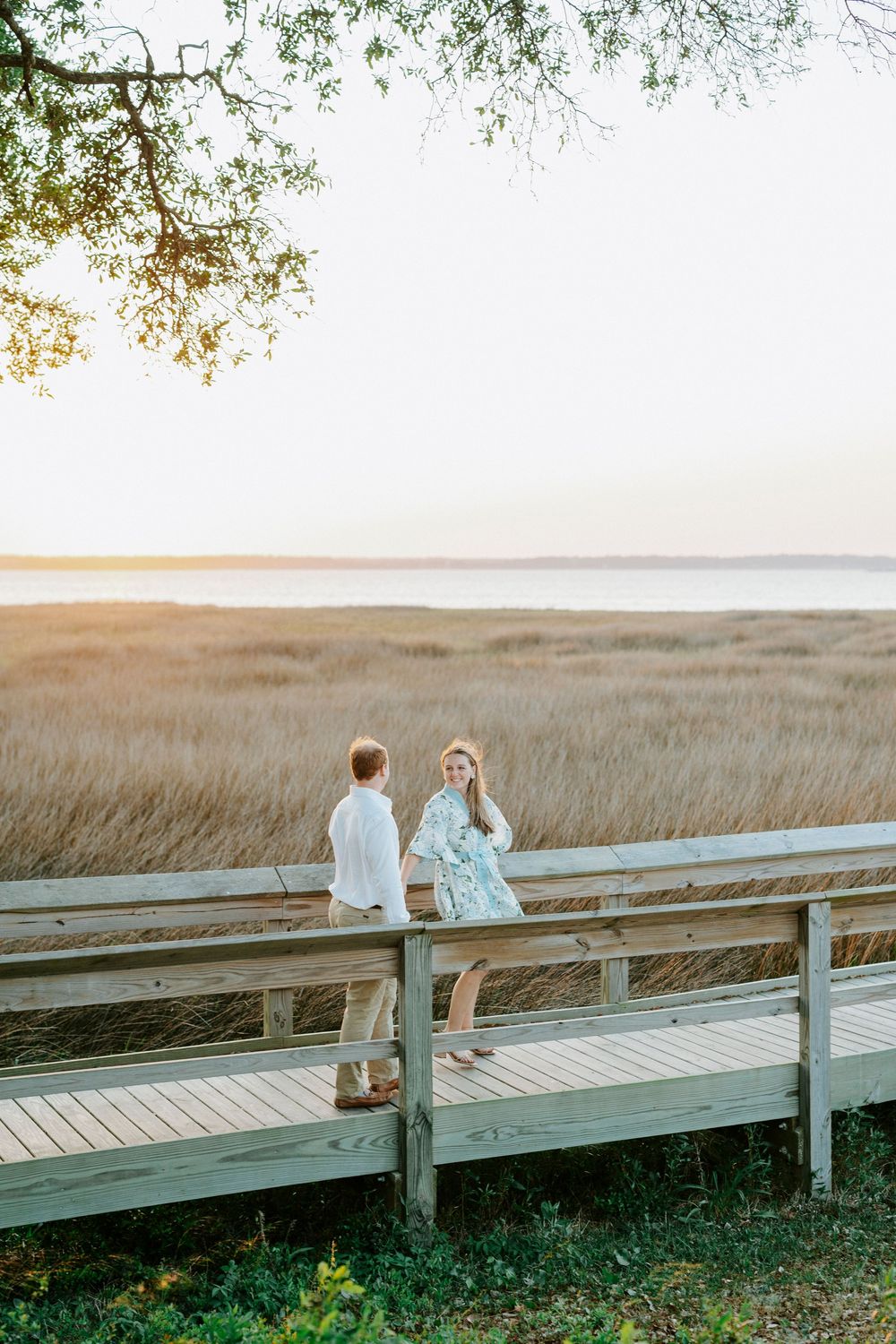 Couple standing on a wooden boardwalk overlooking golden marsh grass and a serene waterway at sunset.