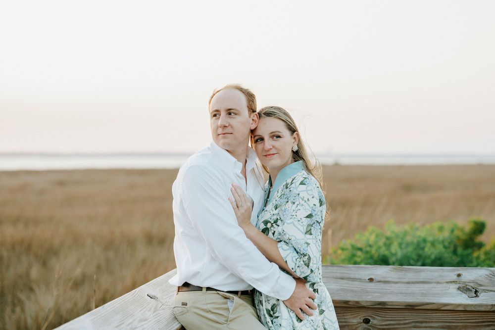 Couple embracing at golden hour on a beach, woman in floral dress and man in white shirt, with marsh grass background.