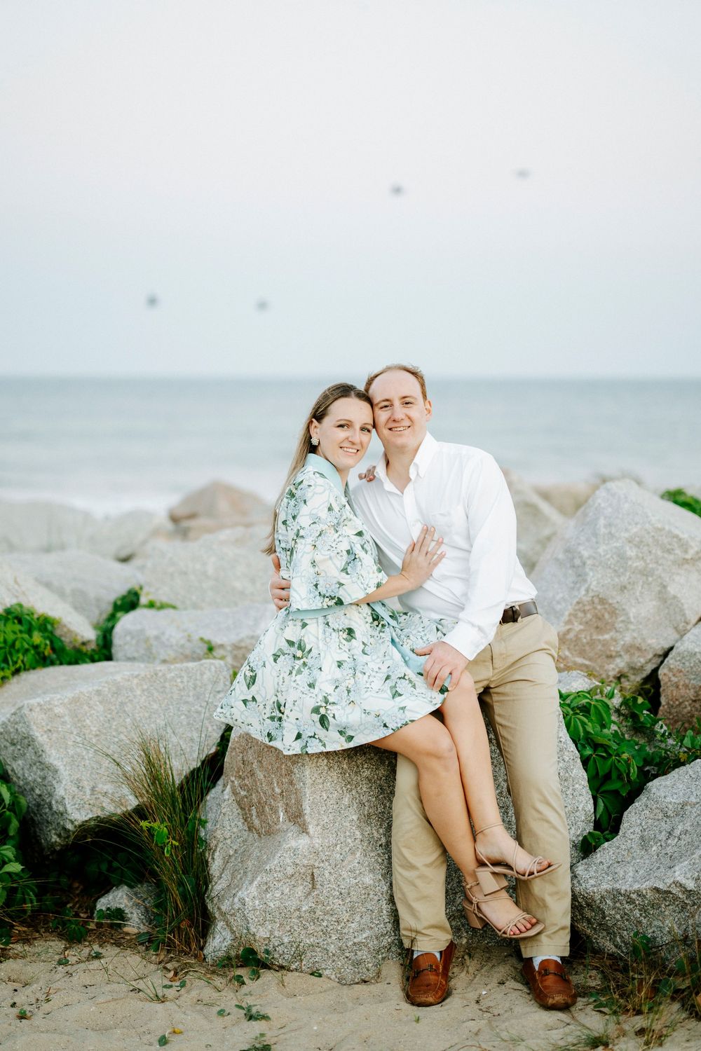Couple sitting on rocks at beach, woman in floral dress embracing man in white shirt, ocean in background.