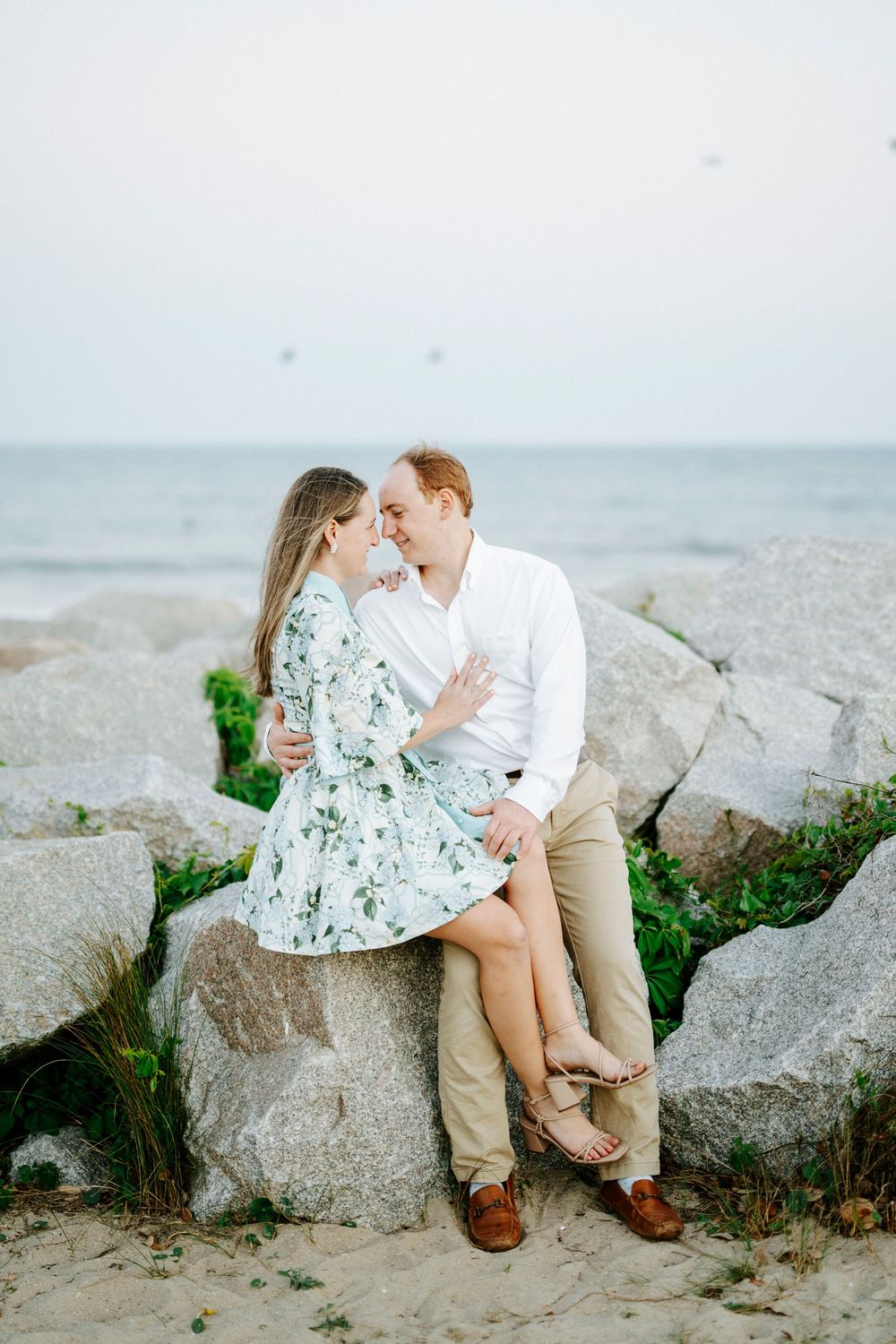 Couple sitting on rocks at beach, woman in floral dress and man in white shirt sharing a romantic moment by the ocean.