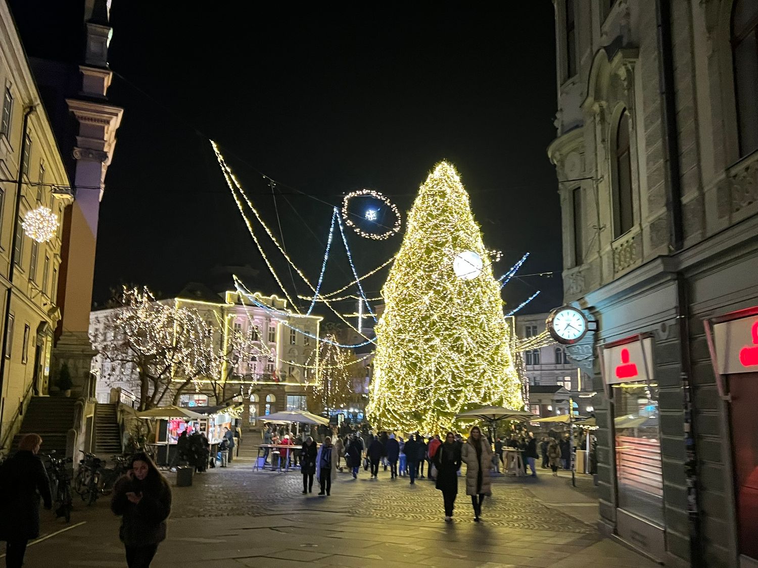 Tall illuminated Christmas tree surrounded by holiday lights and evening shoppers on a festive European pedestrian street at night.