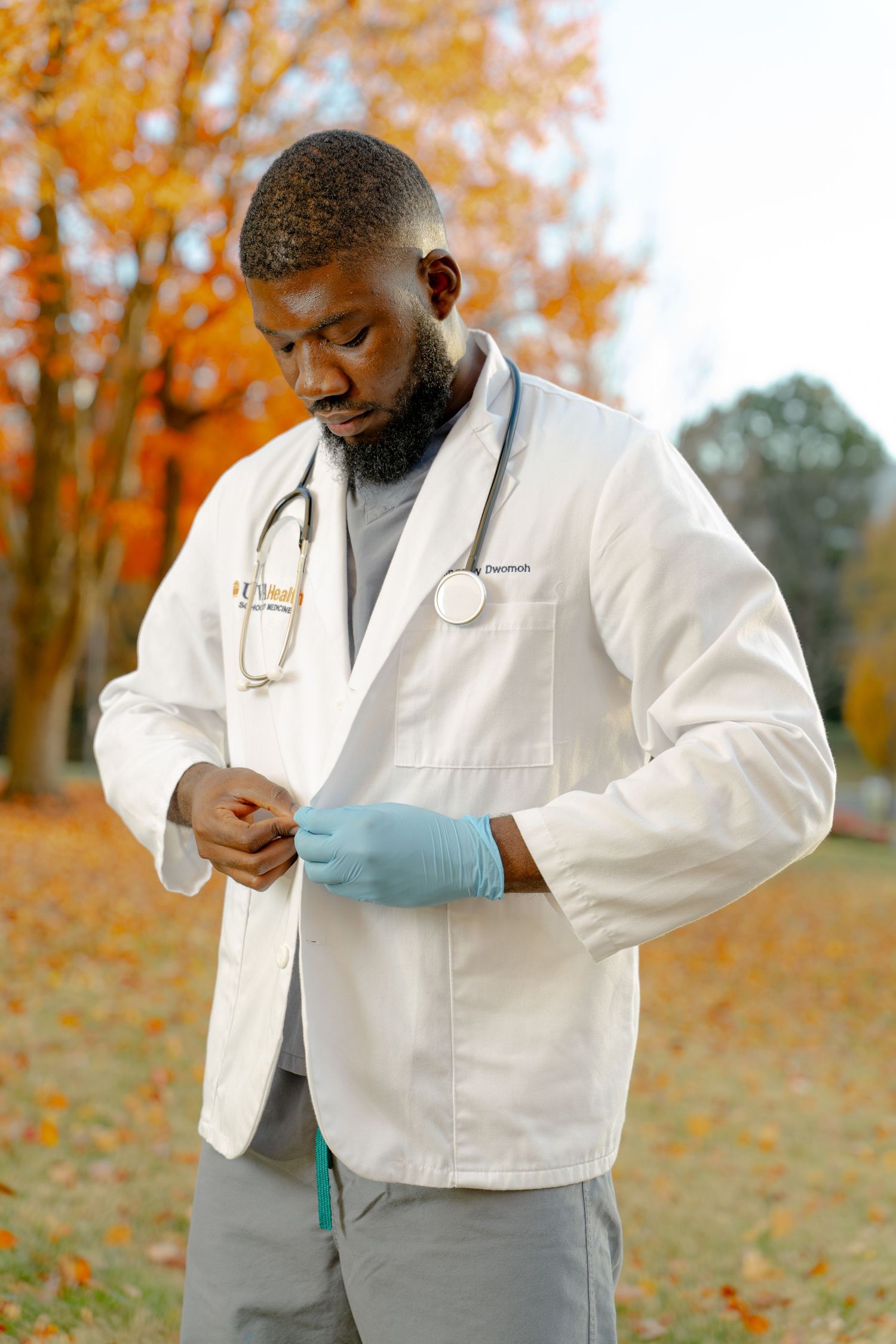 A medical professional in a white coat adjusts their tie against an autumn backdrop with orange foliage.
