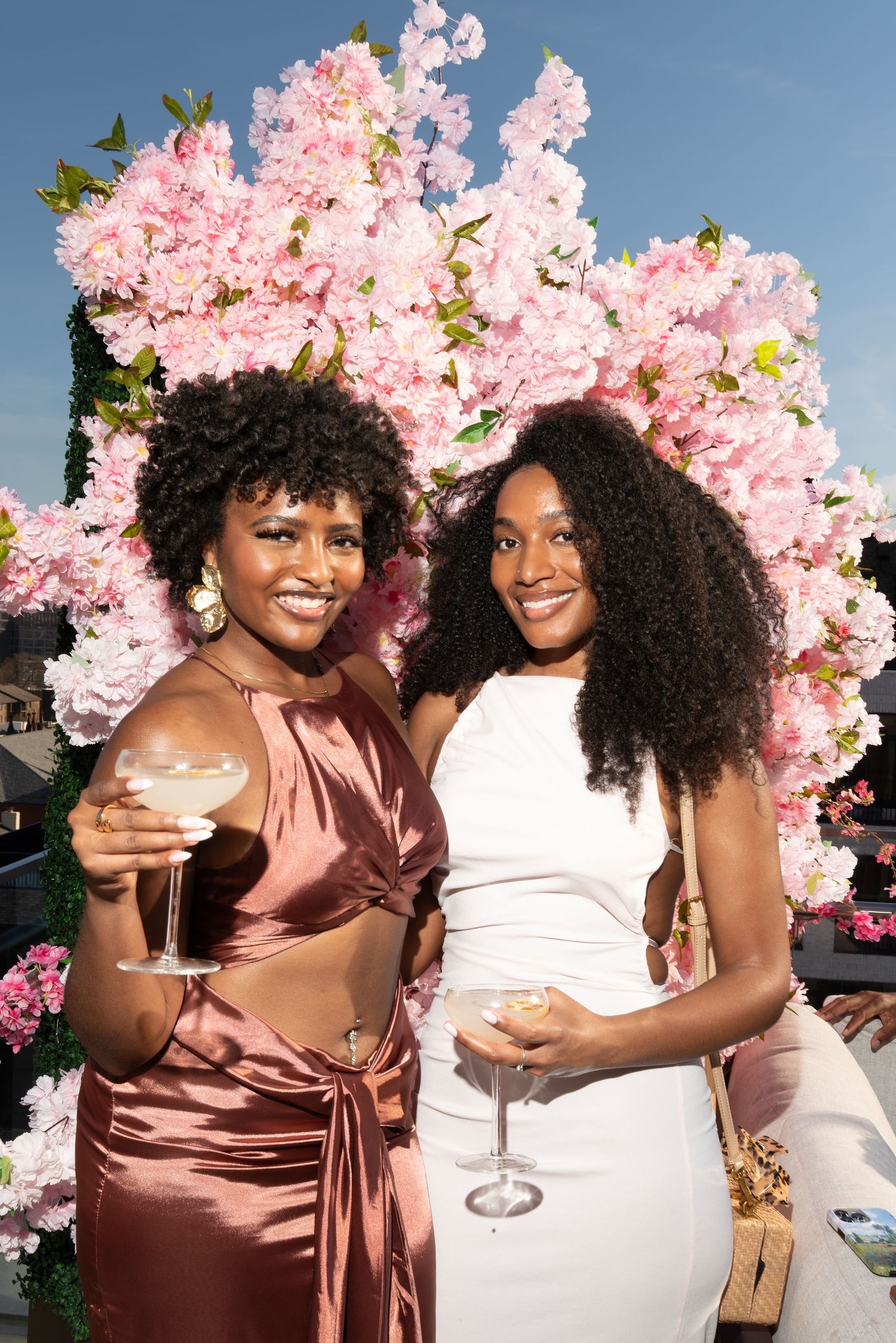 Two friends in satin dresses celebrate with drinks under beautiful pink cherry blossoms at an outdoor event.