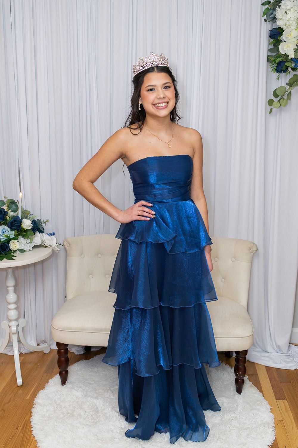 Young woman in navy blue tiered strapless gown wearing a crystal crown, posing elegantly in a decorated studio setting.
