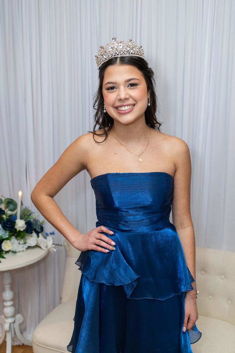 Young woman in blue strapless gown and crystal tiara smiling at formal event, possibly quinceañera or prom.
