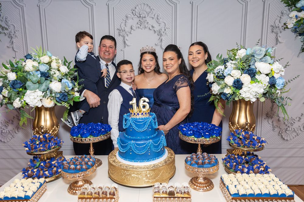 Family posing at a Sweet 16 blue and gold dessert table with floral arrangements.