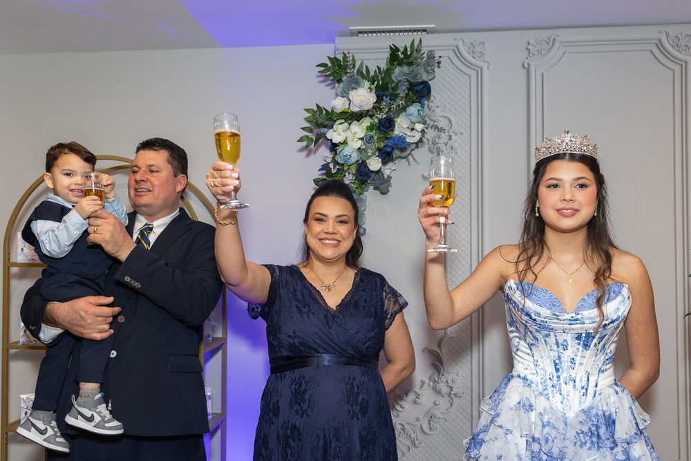 Family celebrating a quinceañera, raising glasses in toast, young girl wearing tiara and blue floral gown at festive party.