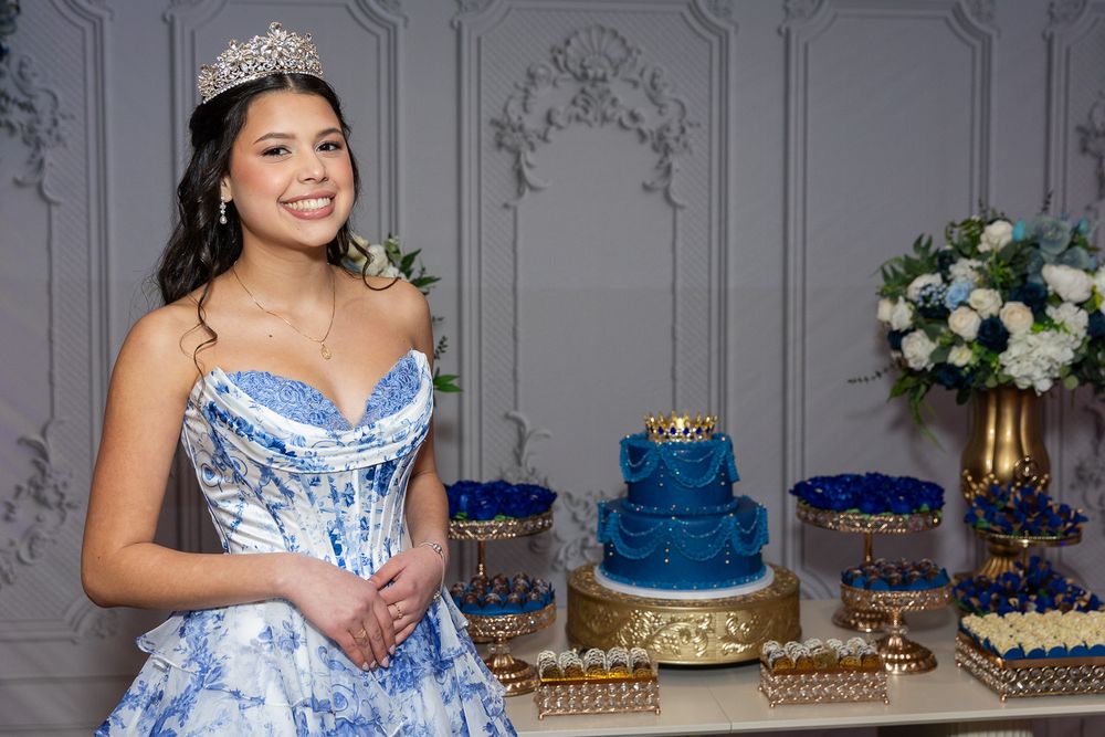Young woman in blue quinceañera gown with tiara poses beside elegant blue cake and floral dessert table.