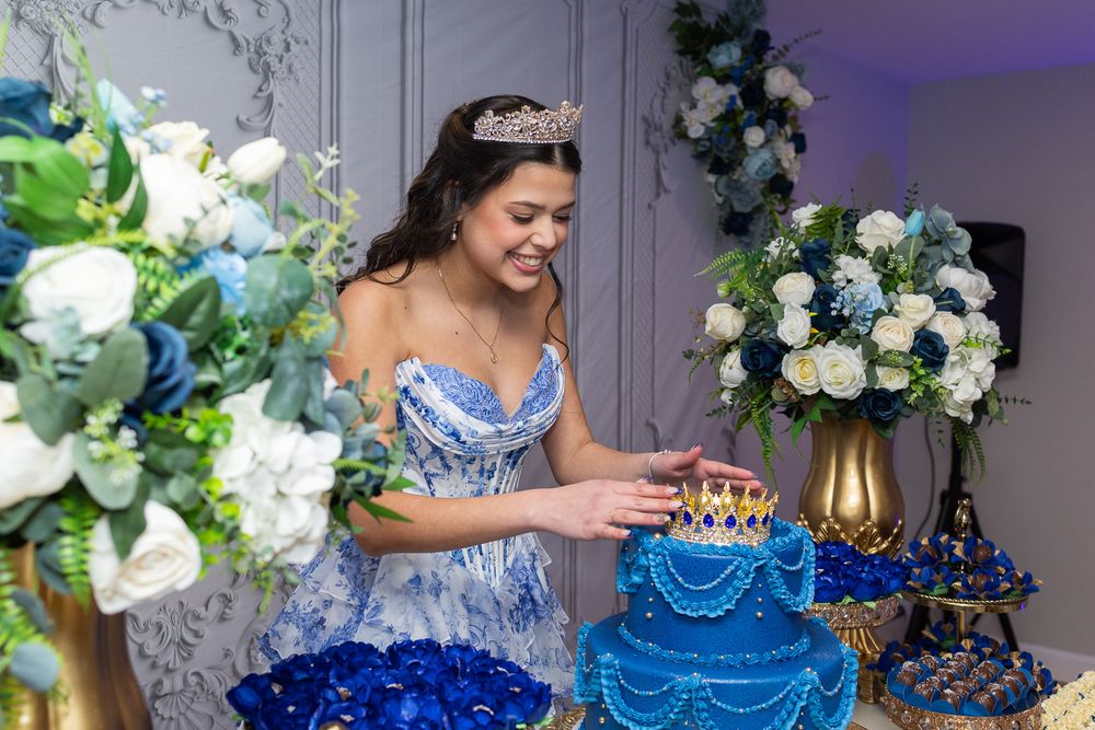 Young woman in blue quinceañera gown and tiara cutting a royal blue tiered birthday cake at elegant celebration.