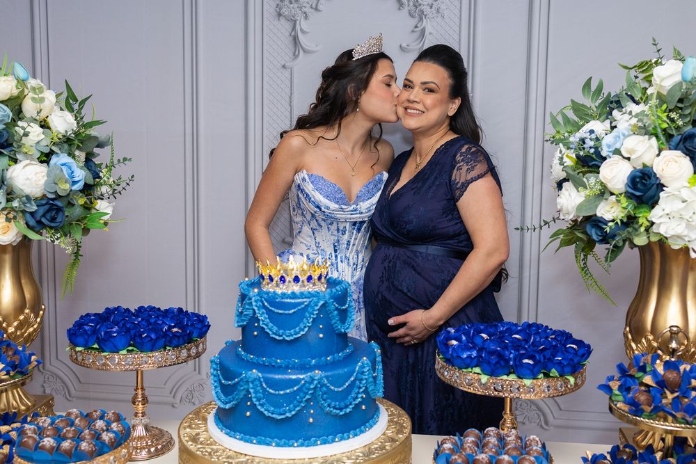 Two women in blue dresses pose by a tiered blue birthday cake surrounded by floral arrangements at a quinceañera celebration.