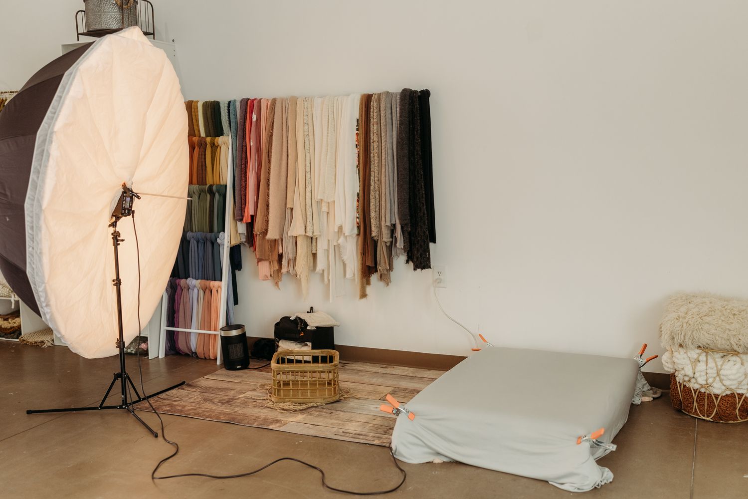 Photography studio setup with colorful fabric backdrops, softbox light, wicker basket, and blanket on wooden floor.