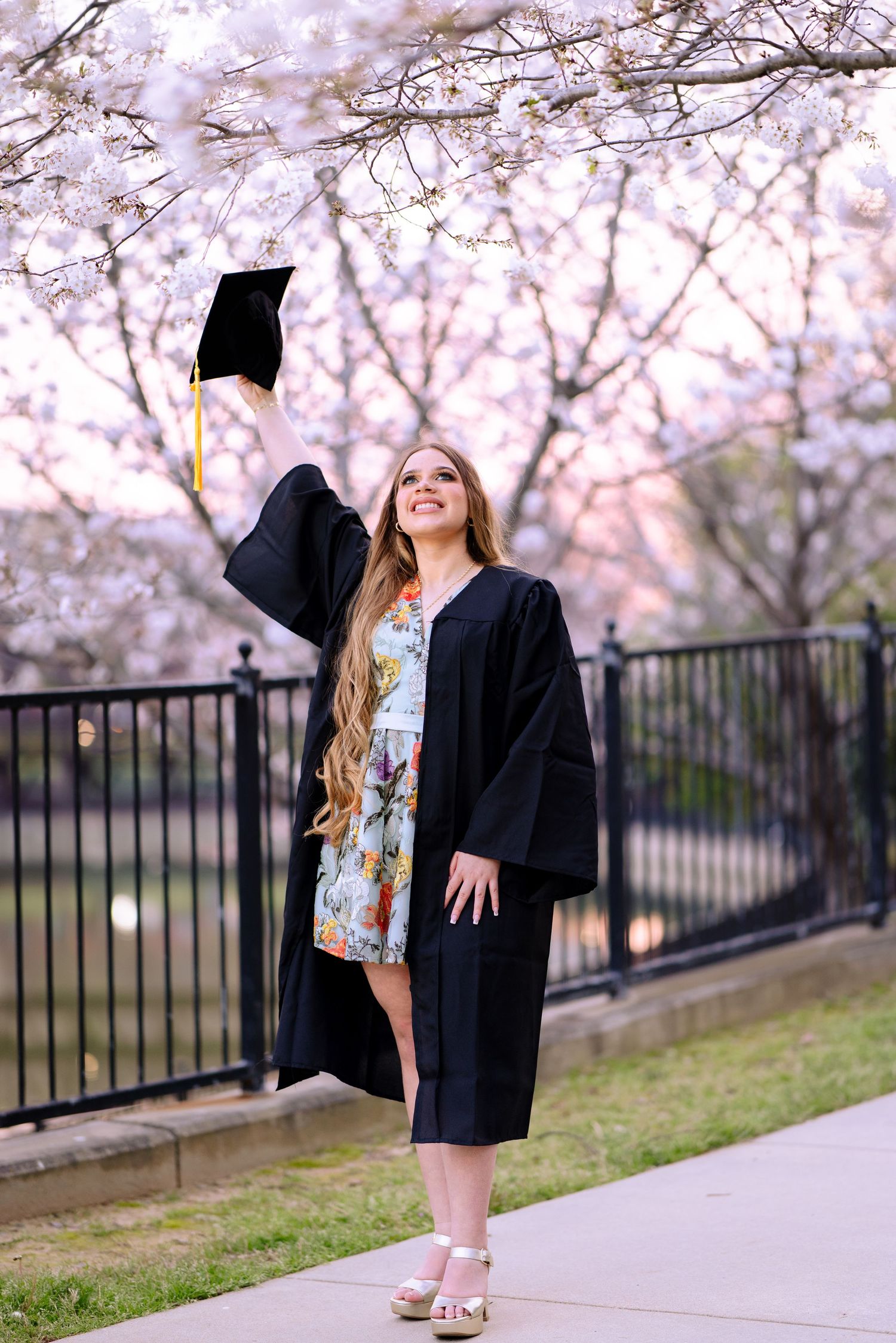Young woman in graduation gown and cap celebrating under cherry blossom trees on a spring day.