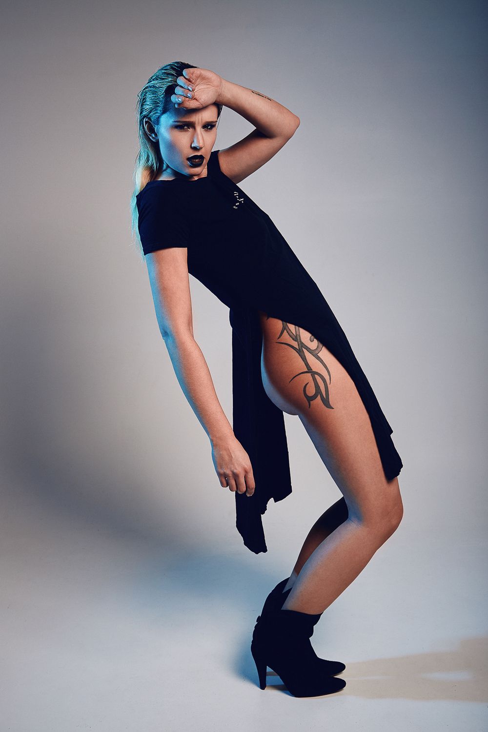 Woman with blue hair in black asymmetric dress with tribal thigh tattoo posing in studio lighting.