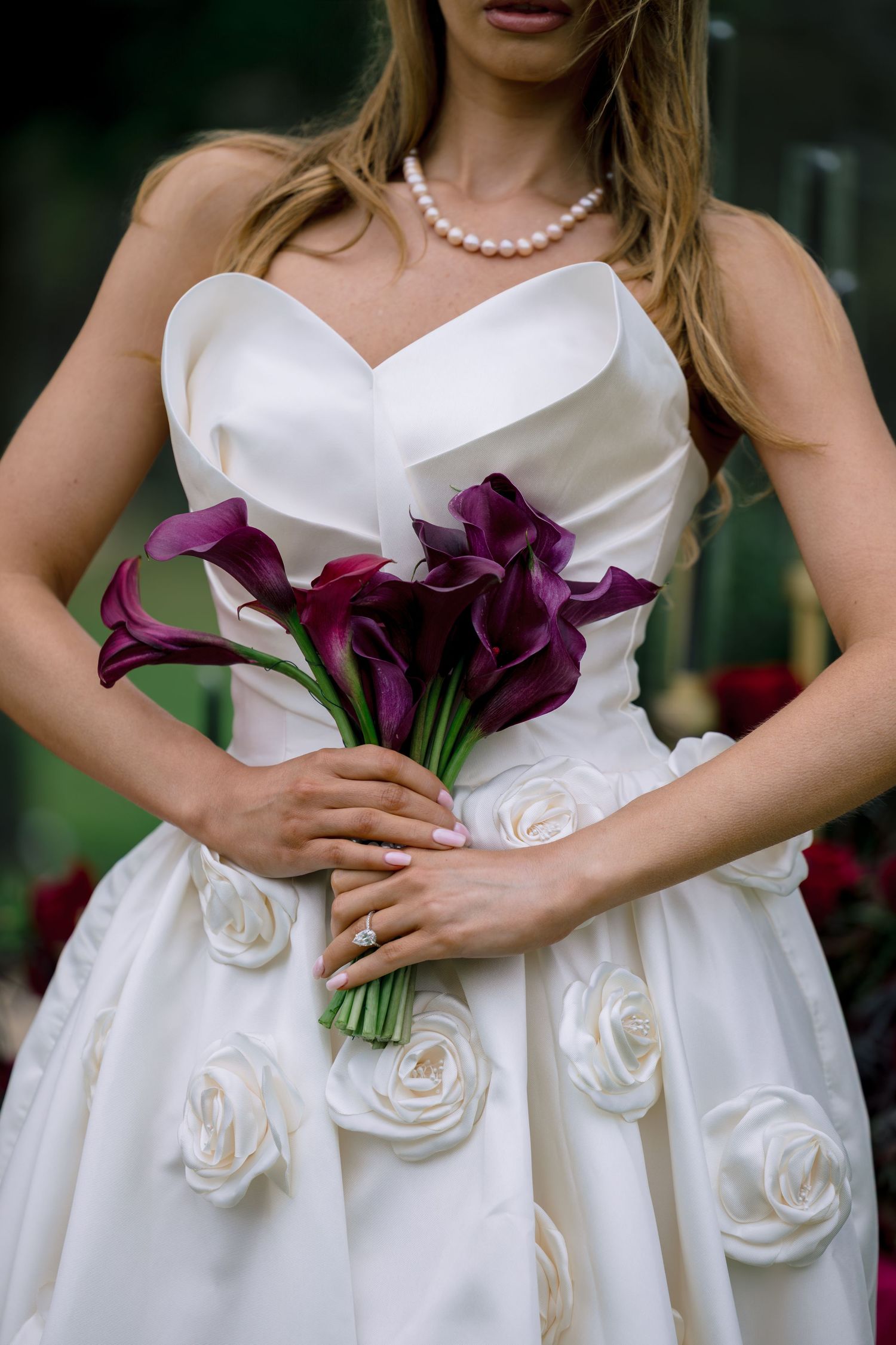 Bride in strapless white gown with rose appliques holds deep purple calla lily bouquet, wearing pearl necklace.