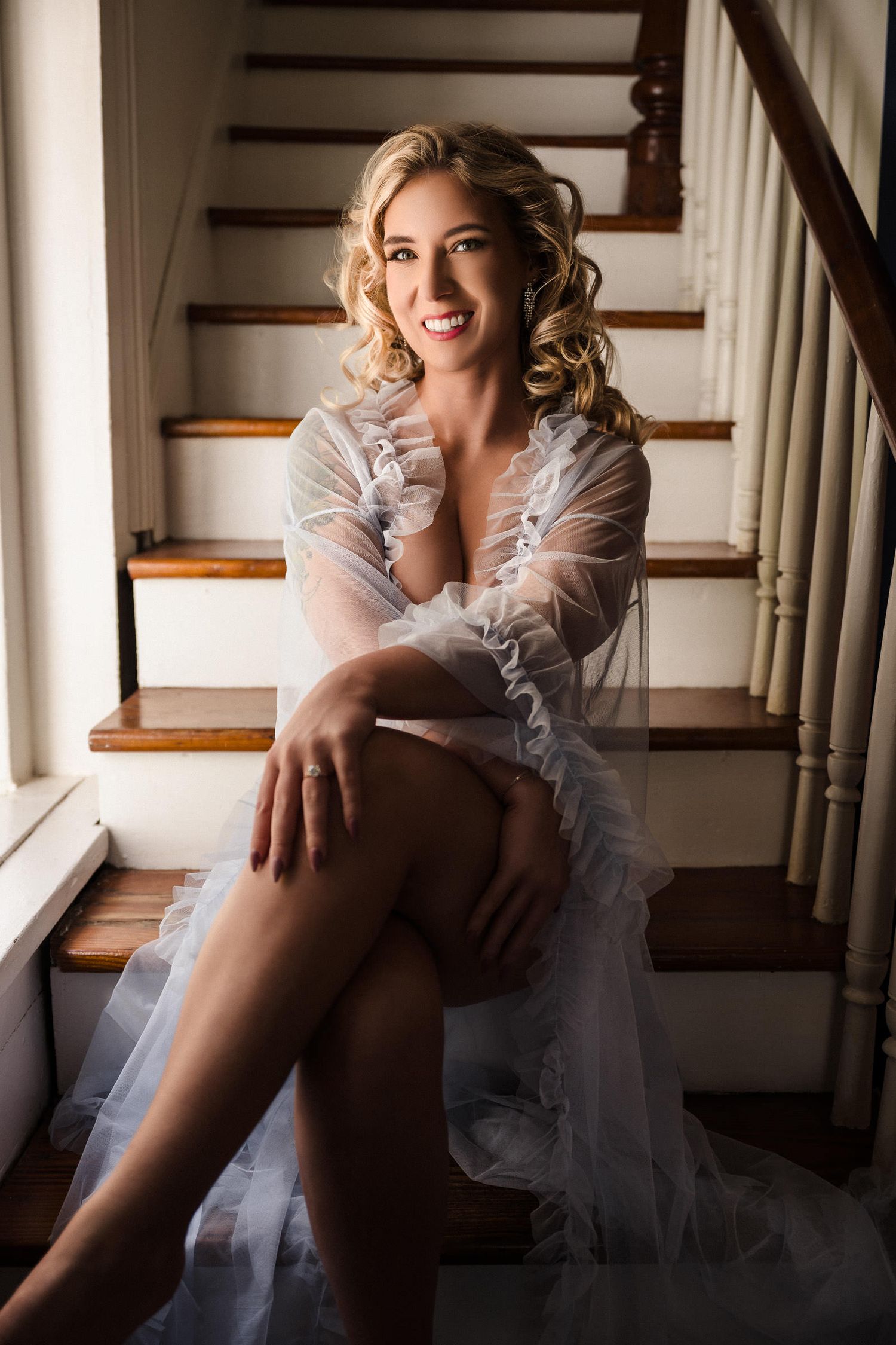 Bridal boudoir photography New Orleans - Smiling blonde woman in sheer white robe sitting on wooden staircase, posing elegantly.