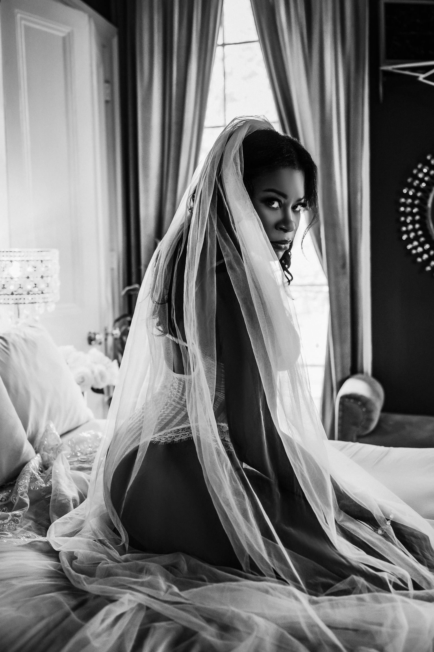 Bridal boudoir photography New Orleans -  Black and white photo of a bride in lingerie draped in a flowing veil, sitting on a bed.