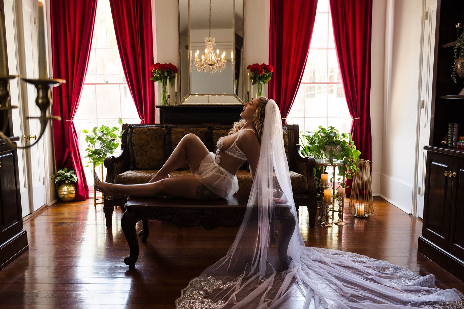 Bridal boudoir photography New Orleans -  Bride in lingerie and veil reclines on a chaise lounge in an elegant room with red curtains.