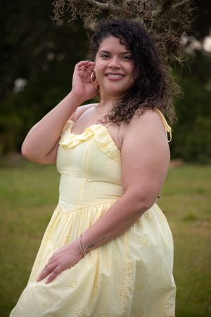 A smiling woman in a yellow eyelet sundress poses outdoors near a tree in a lush green field.