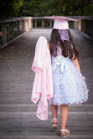 Young girl in blue dress and pink graduation cap walks on wooden bridge holding pink gown.