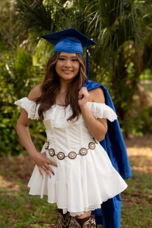 Young woman in white dress and blue graduation cap and gown smiling outdoors with tropical greenery background.