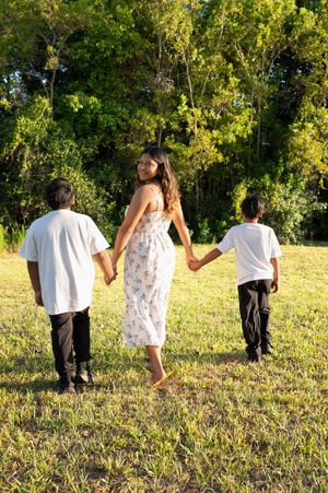 A woman in a floral dress holds hands with two children walking barefoot across a sunny green field toward trees.