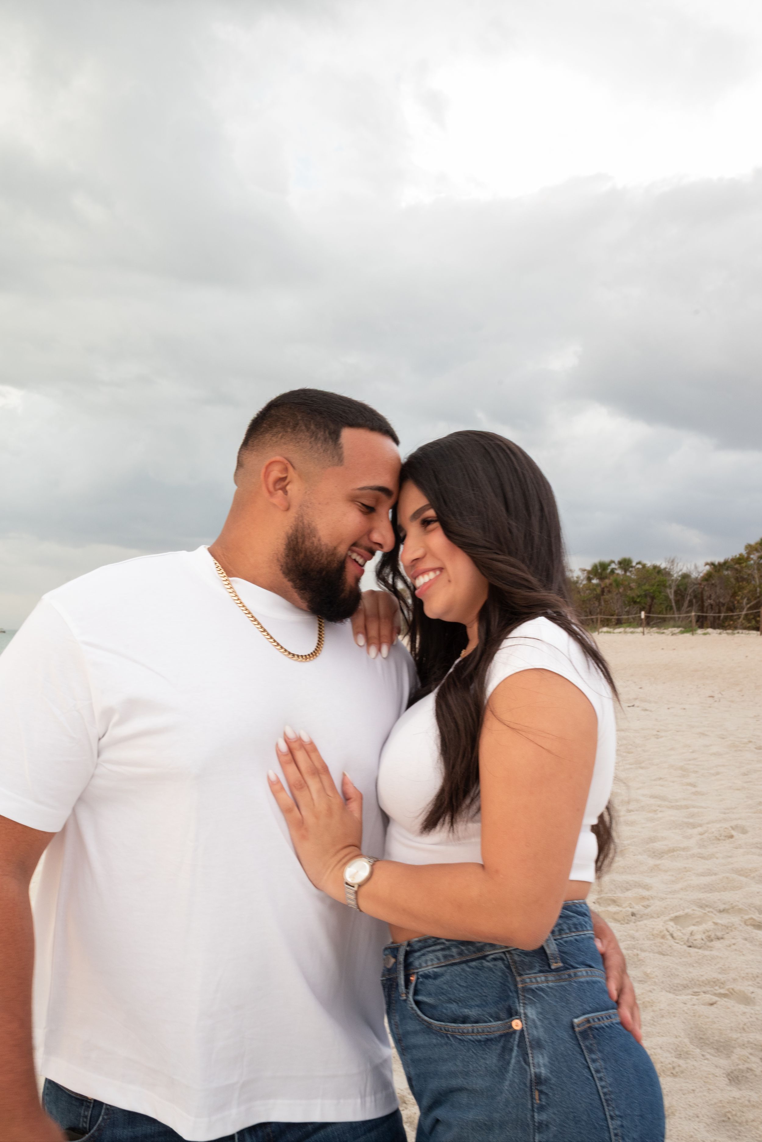 Couple in matching white t-shirts and jeans share a romantic moment on a cloudy beach.