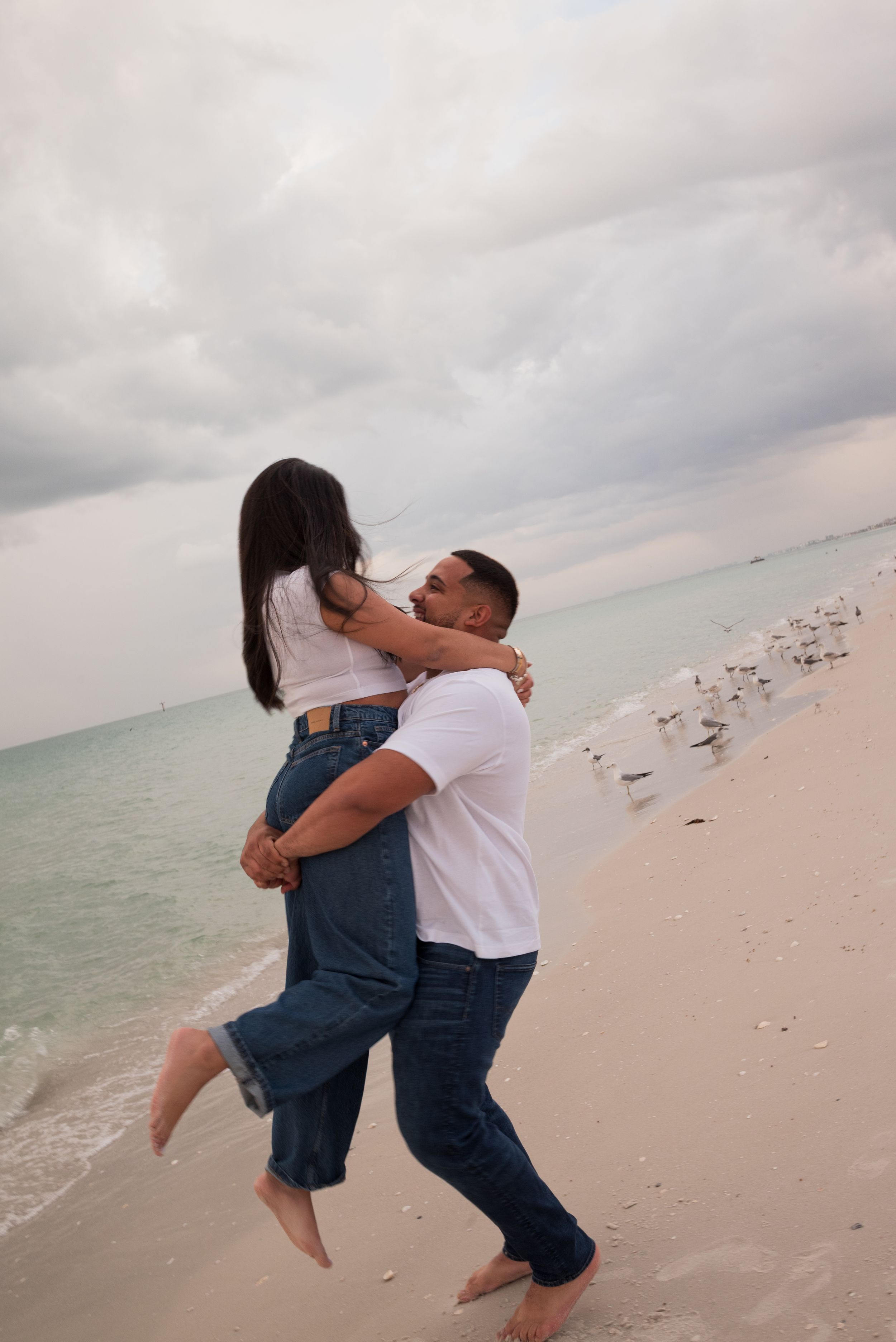 A couple embraces on a white sand beach, the man lifting the woman as waves and cloudy skies set a romantic scene.