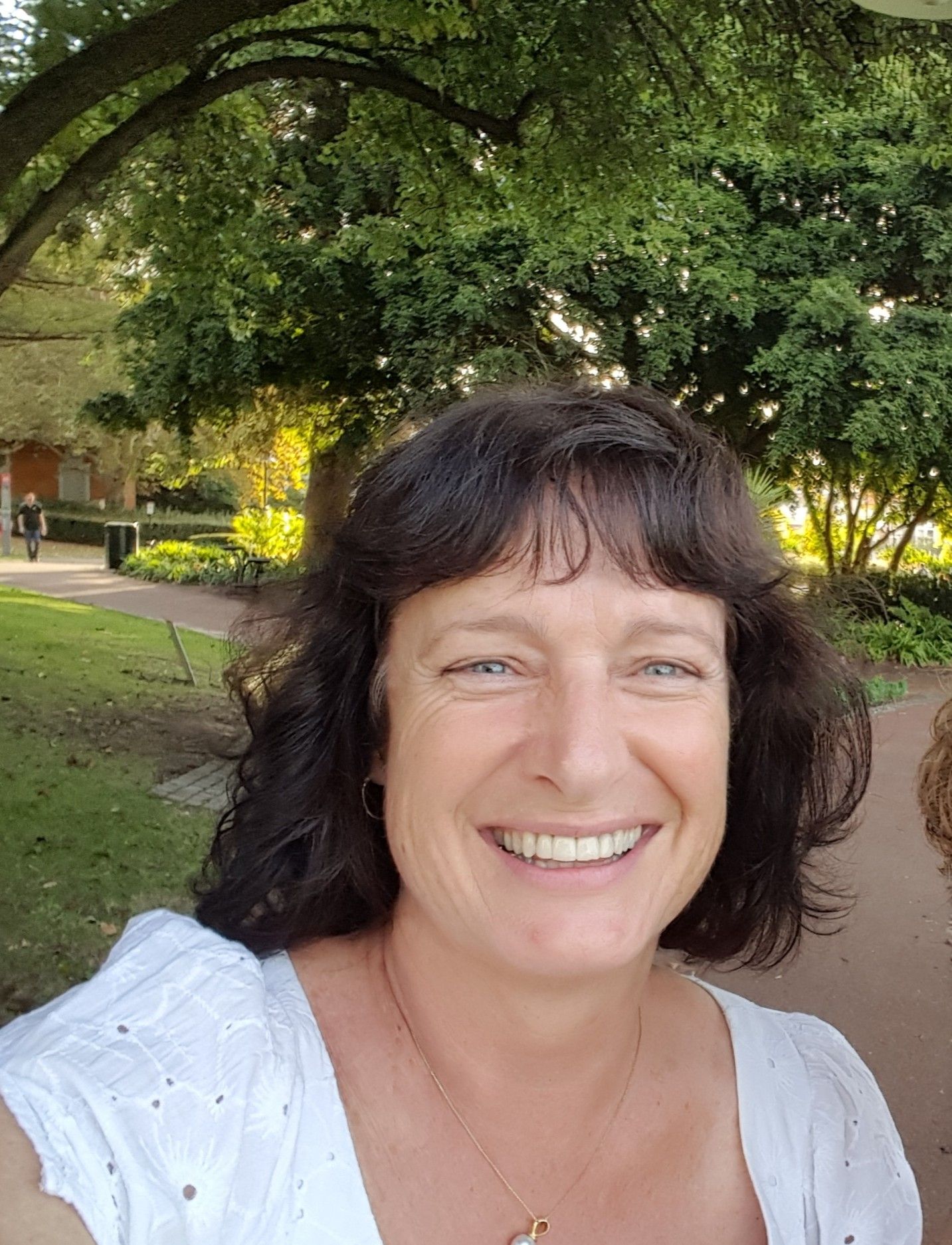 Smiling woman with dark curly hair outdoors in a sunny park with green trees in the background.