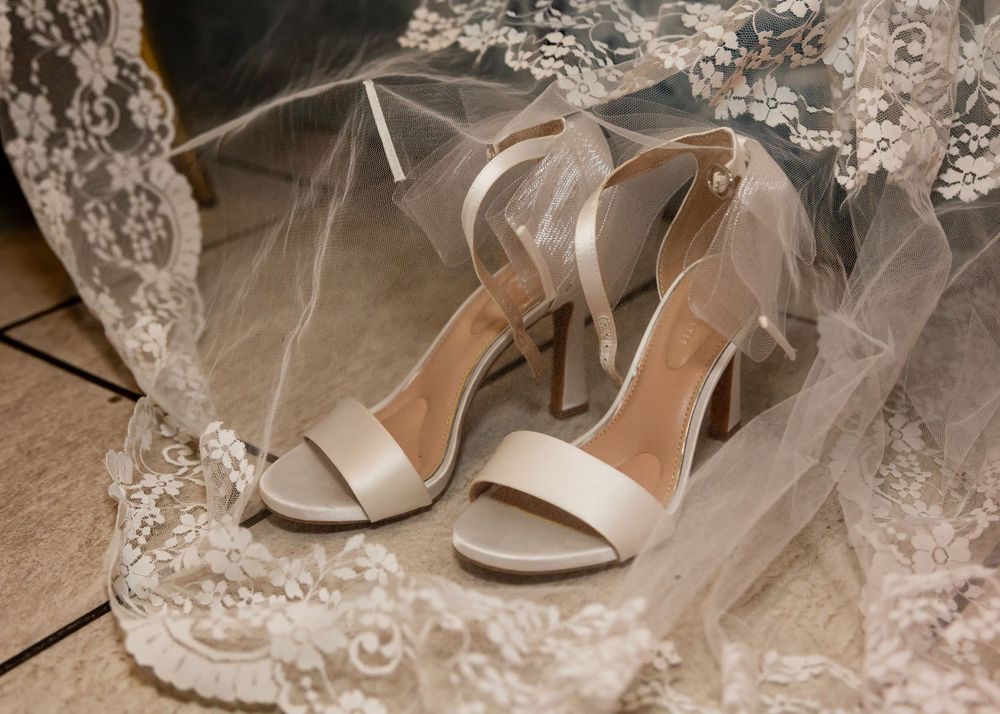 Elegant ivory bridal heels with ankle strap resting on lace wedding veil on wooden floor.