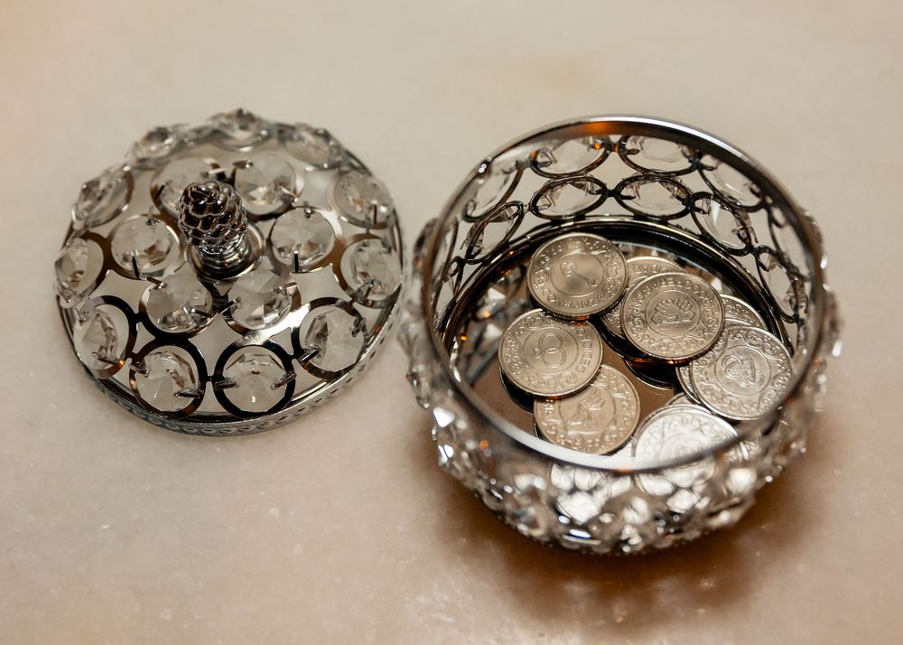 Silver crystal decorative trinket box with ornate circular patterns, lid ajar, filled with assorted coins on beige surface.