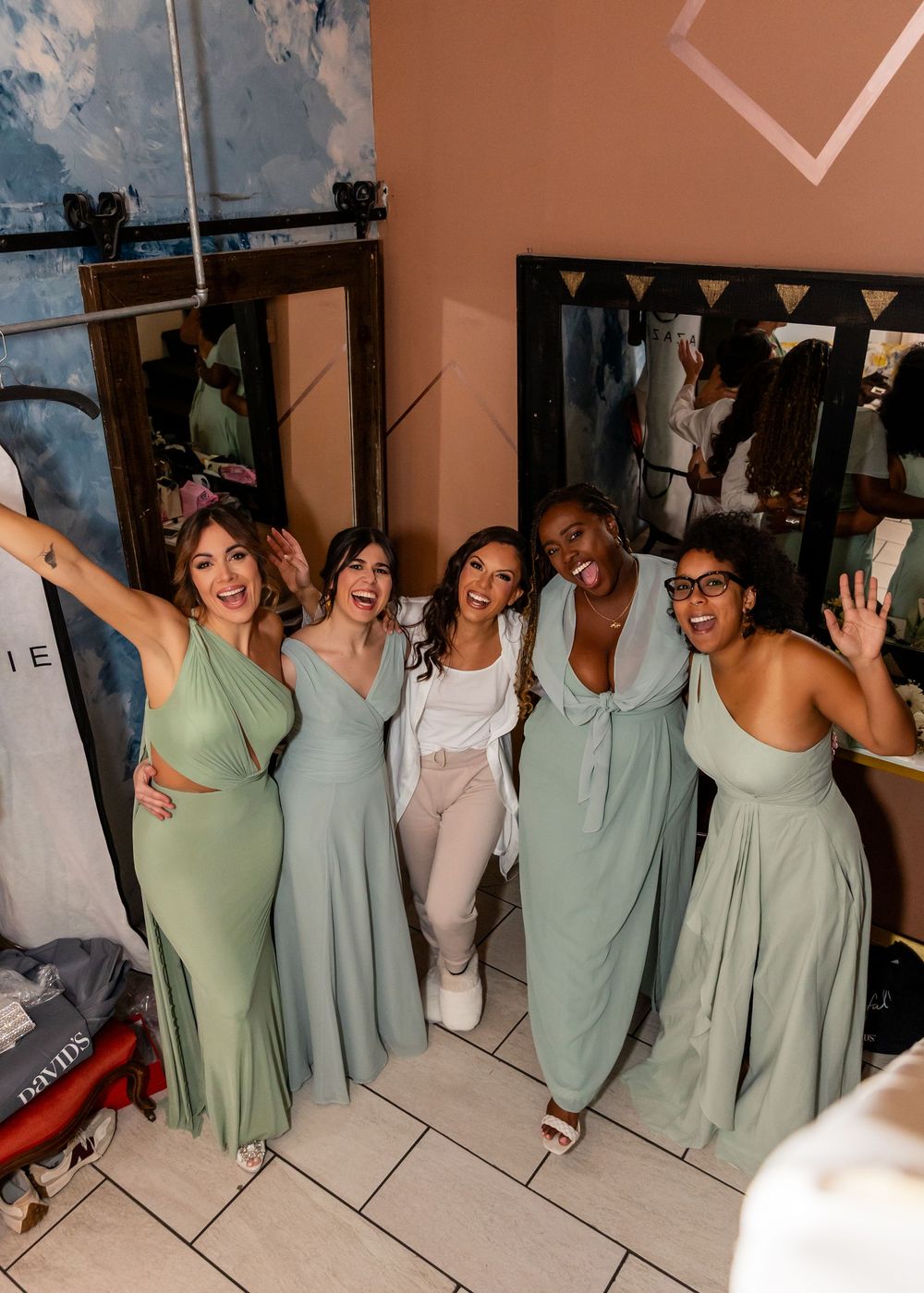Five bridesmaids in sage green dresses celebrate joyfully together in bridal suite before wedding.