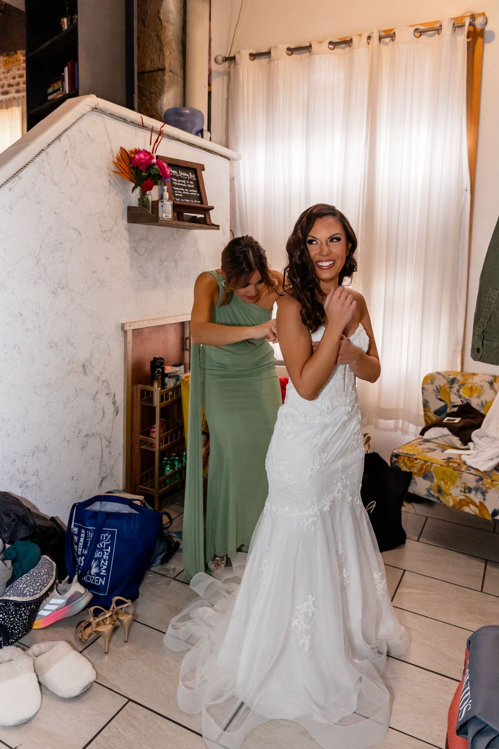 Smiling bride in white mermaid gown gets dress fastened by bridesmaid in sage green dress in a bridal getting-ready room.