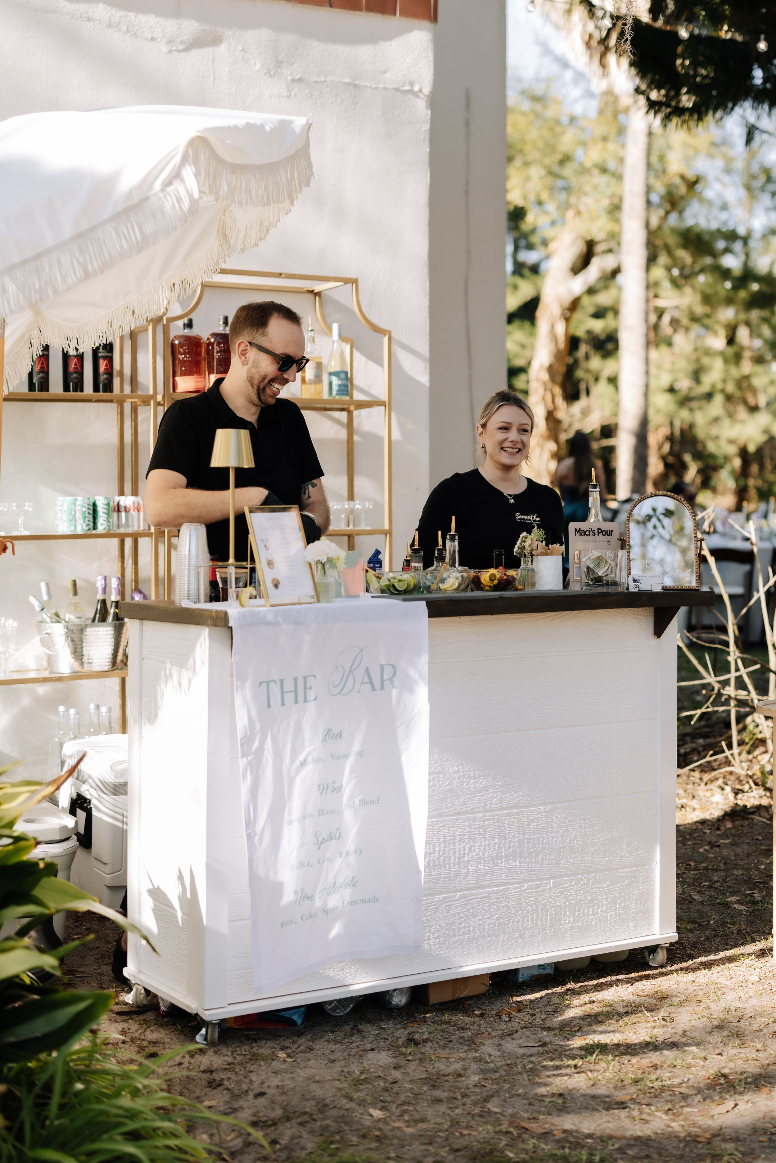 Two smiling bartenders stand behind a white mobile bar cart at an outdoor event under a white canopy.
