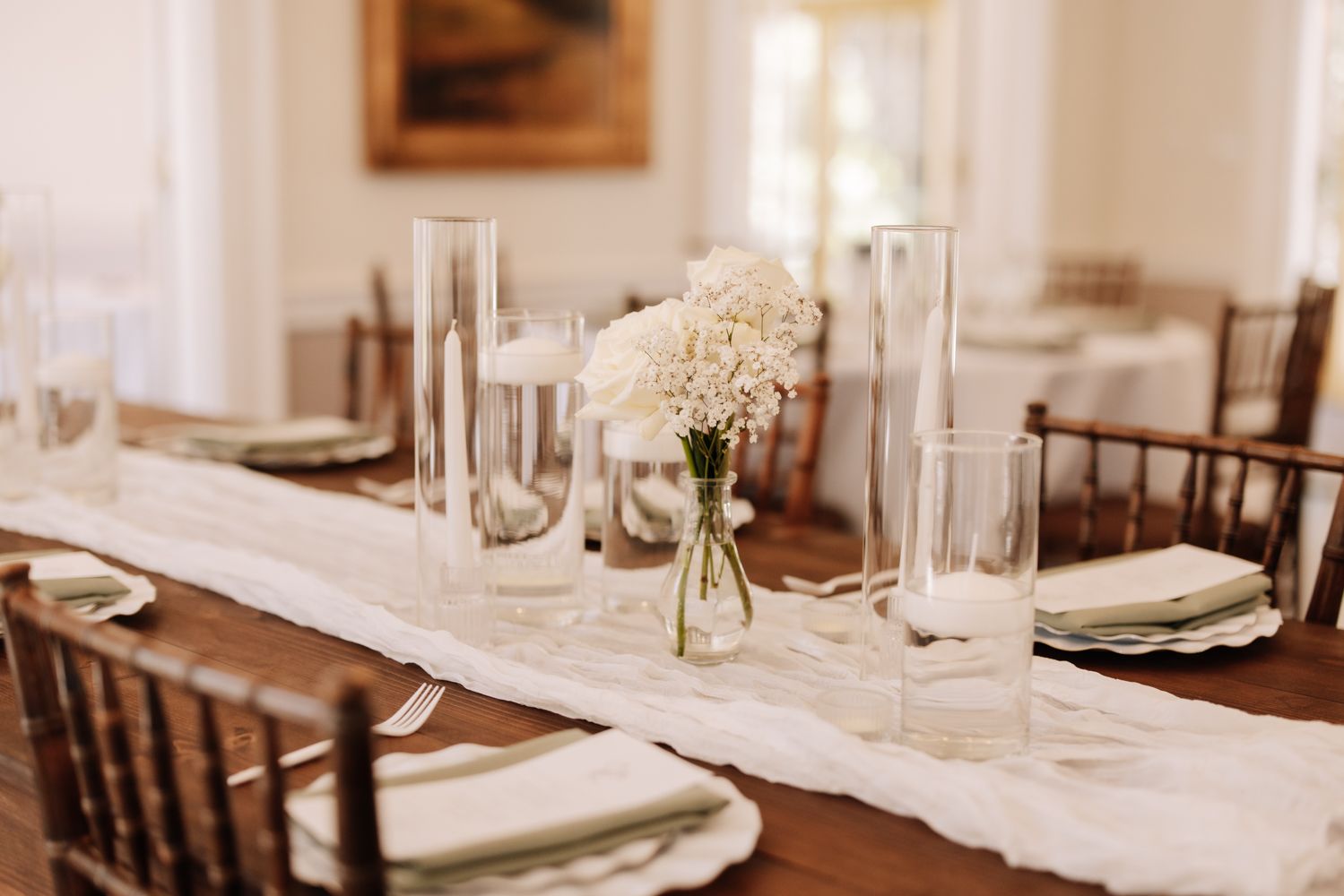 Elegant wedding reception table with white linen runner, glass cylinder vases, and white floral centerpieces on a wooden table.
