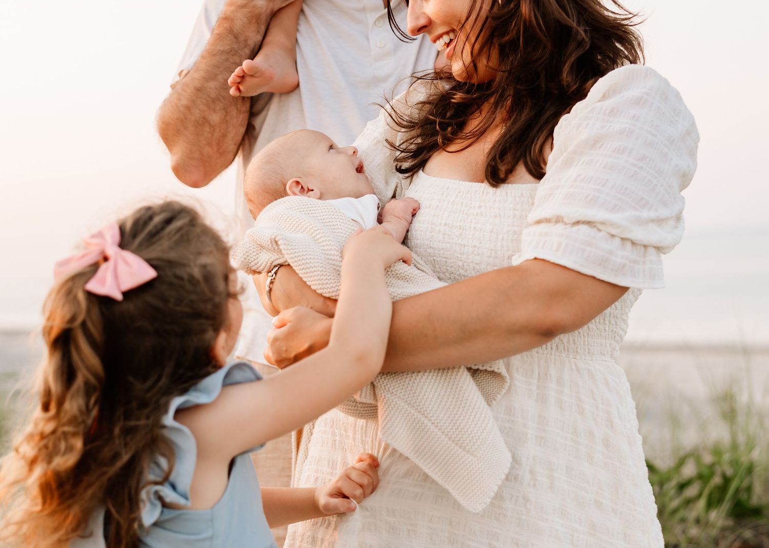 Family photo of parents holding infant and toddler girl in pink bow outdoors at golden hour.