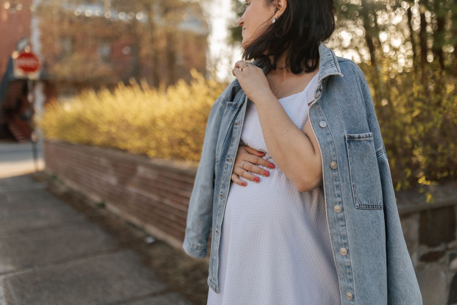 Pregnant woman in white dress and denim jacket holding baby bump outdoors on sunny day.