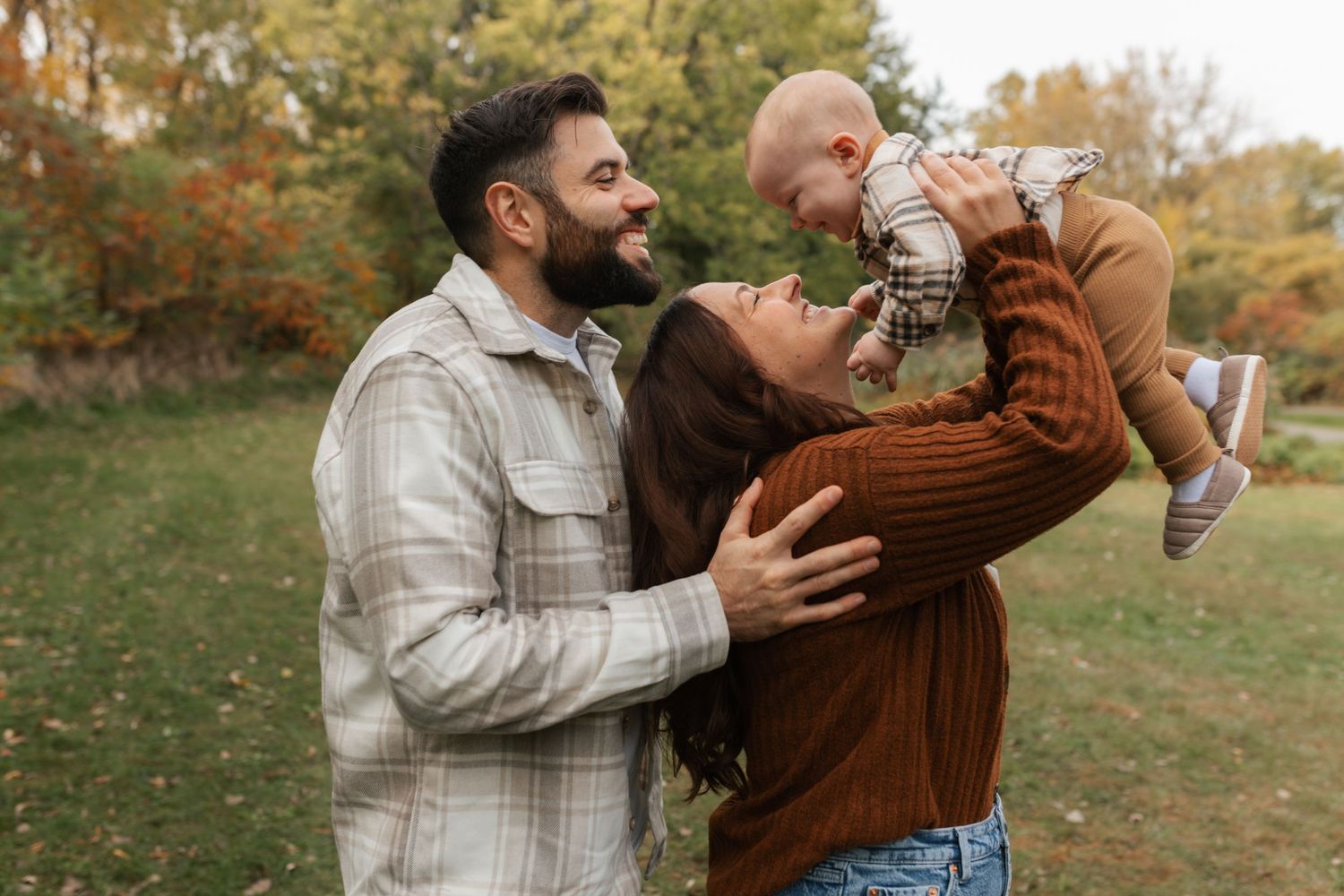 Happy family with parents lifting smiling baby outdoors in autumn park with colorful fall foliage.
