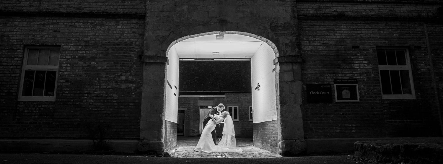 Black and white photo of a bride and groom sharing a romantic kiss in an arched doorway of a rustic brick building.