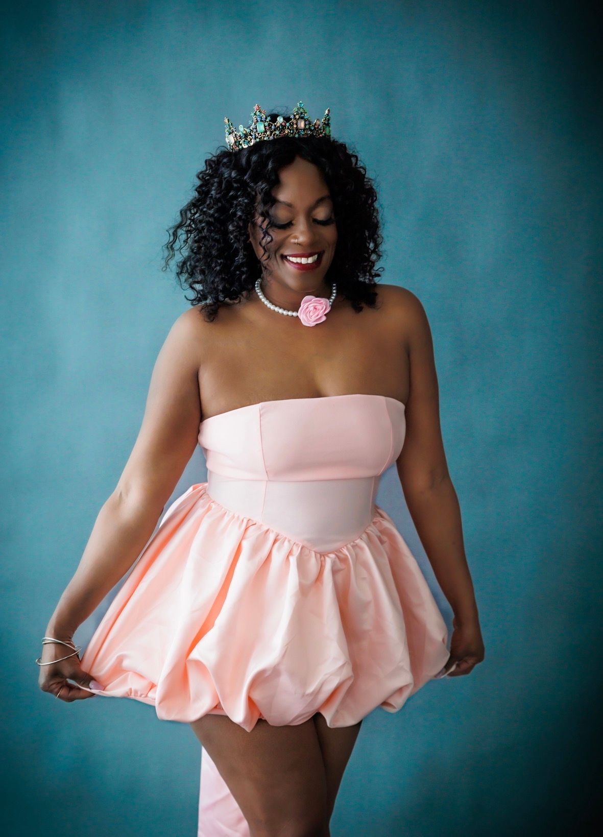 A smiling woman in a pink bubble skirt dress and crown poses against a teal background.