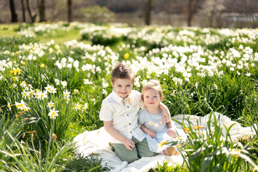 Two smiling siblings sitting on a blanket in a lush field of white spring daffodils.