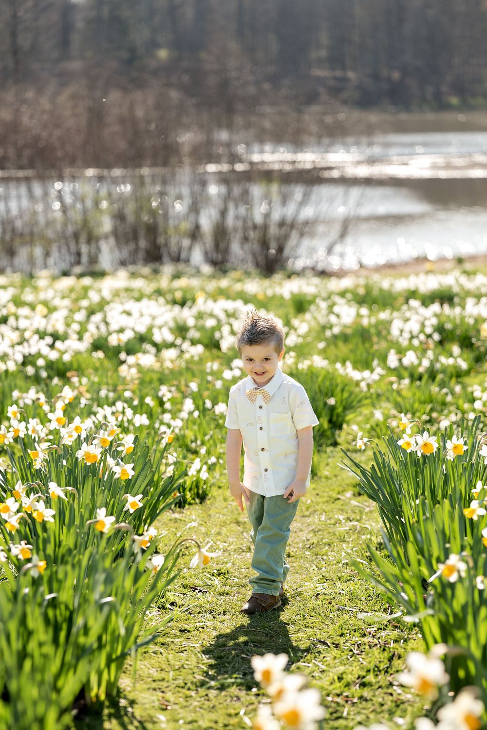 Young boy in a white button-up shirt stands smiling in a lush field of white and yellow daffodils.