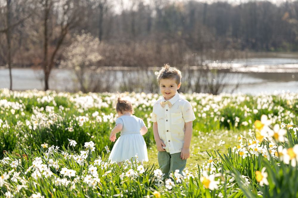 Young boy in white shirt holds hands with toddler girl in blue dress among white spring daffodils.