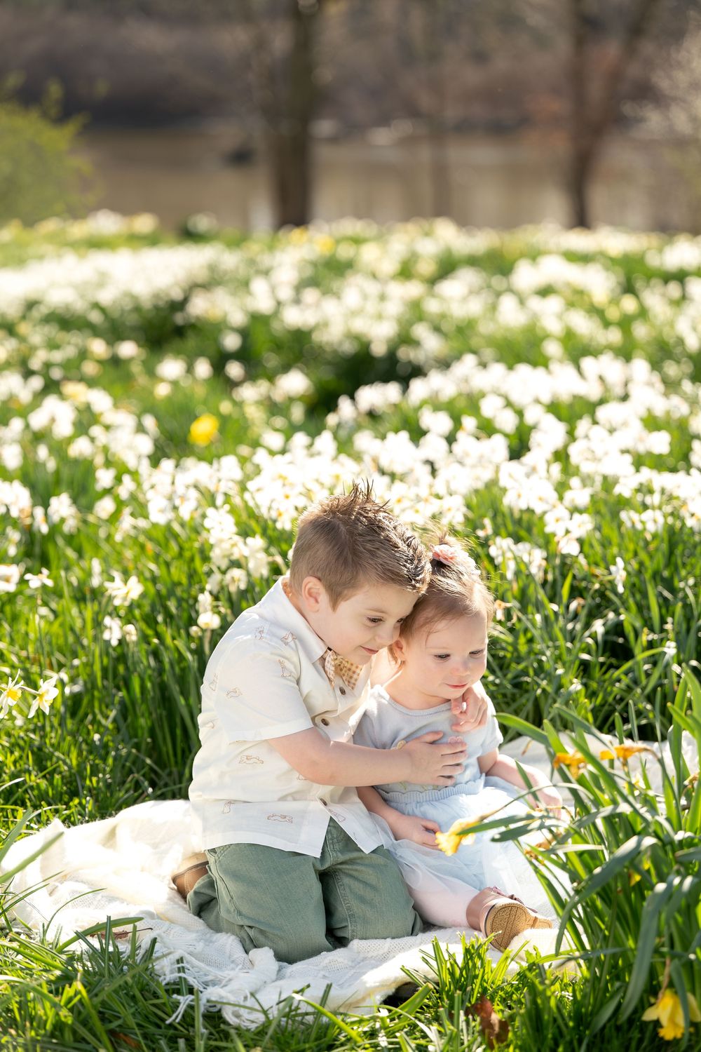 Boy and toddler girl sitting together on a blanket surrounded by blooming white daffodils.