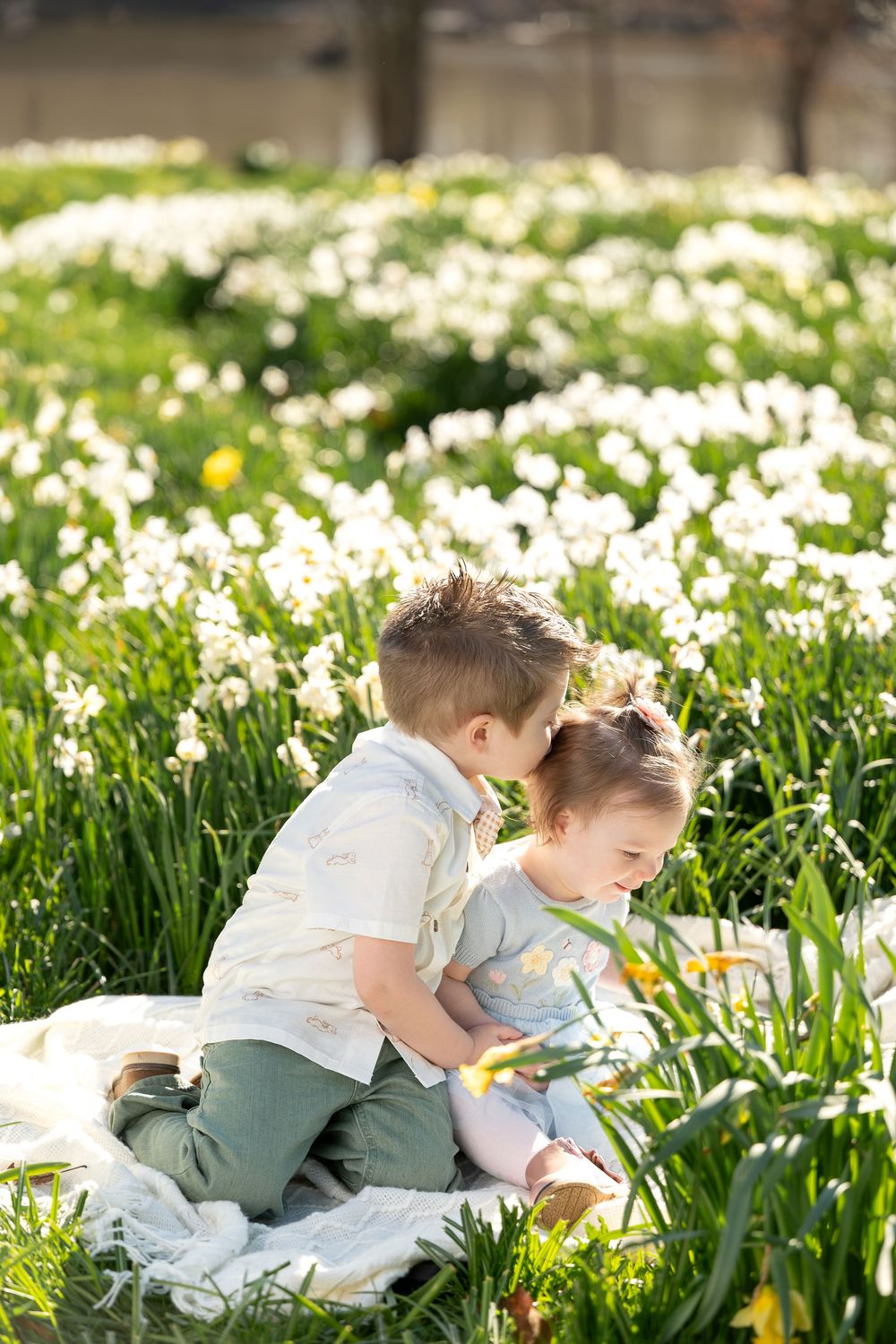 Two young children sitting close together on a blanket amid a field of white daffodils.