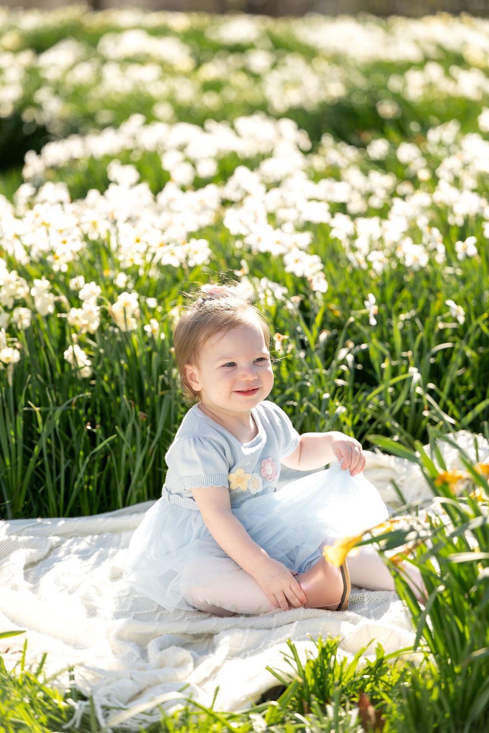 Cheerful baby girl in light blue dress smiling while sitting in a sunny daffodil meadow.