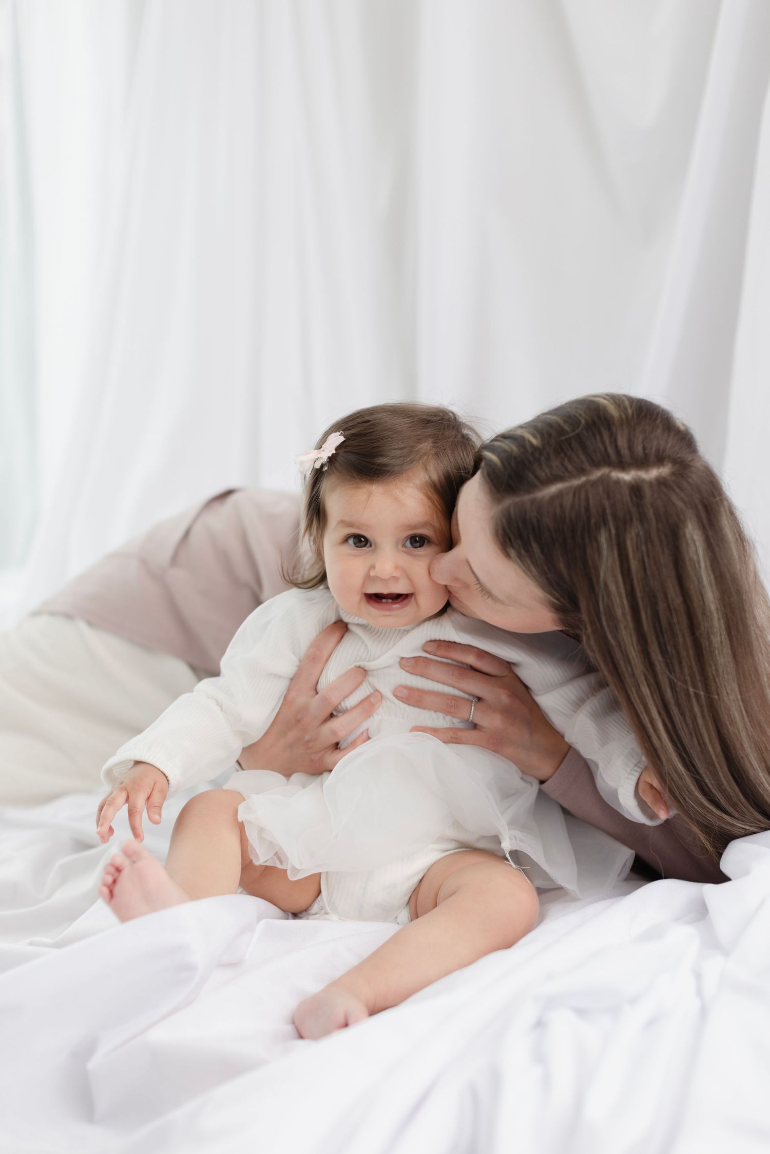 Mother tenderly kissing happy baby girl in white outfit on soft white bedding. Elizabeth Lang Photography Surrey