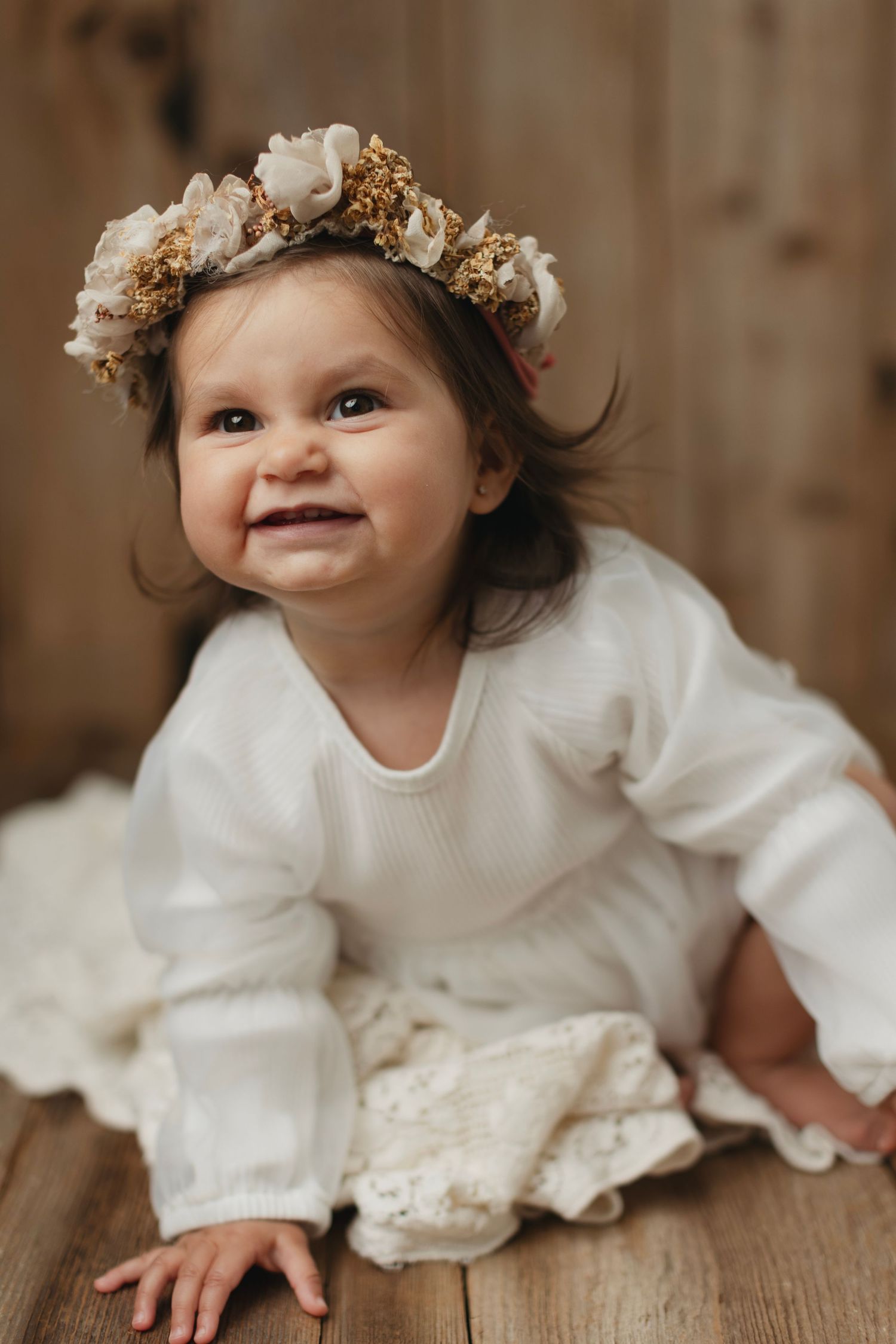 Smiling baby girl in white knit dress and floral crown sitting on wooden floor. Elizabeth Lang Photography Surrey