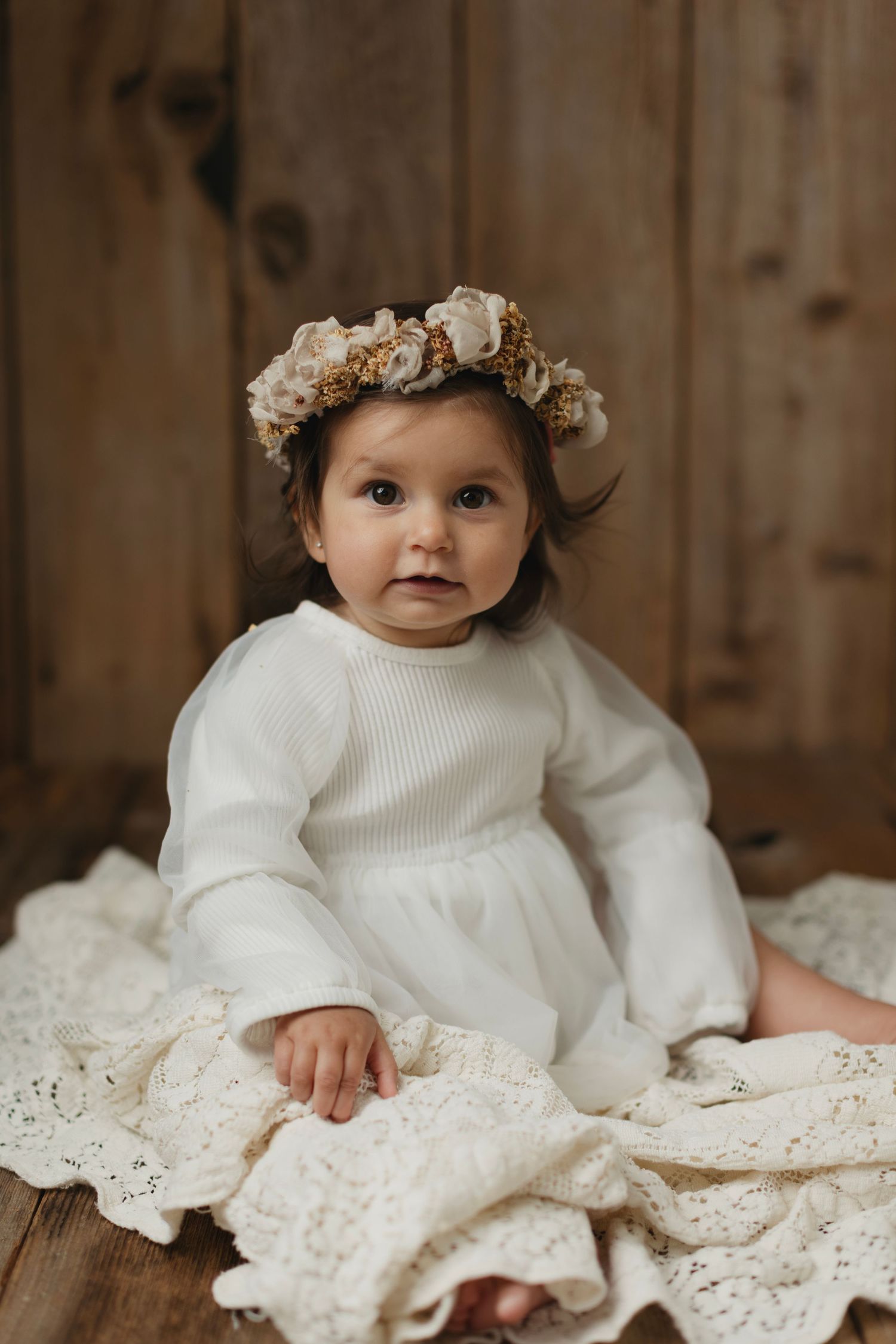 Baby girl in white ribbed dress and floral crown sits on lace blanket against rustic wood backdrop. Elizabeth Lang Photography Surrey